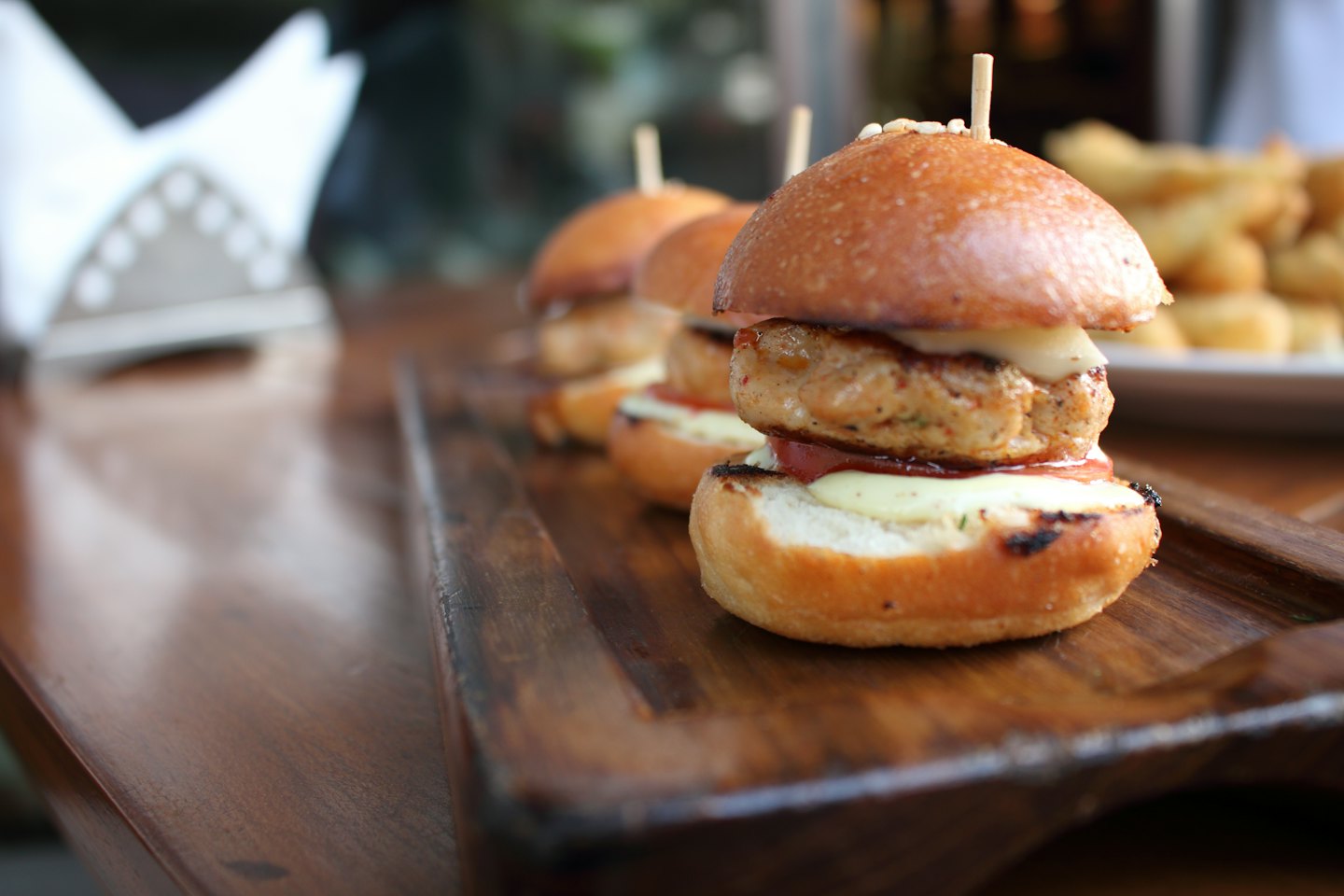 Three mini chicken burgers with cheese and tomato on a wooden tray.