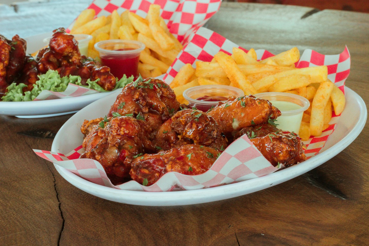 Two plates of glazed chicken wings and fries on a wooden table.