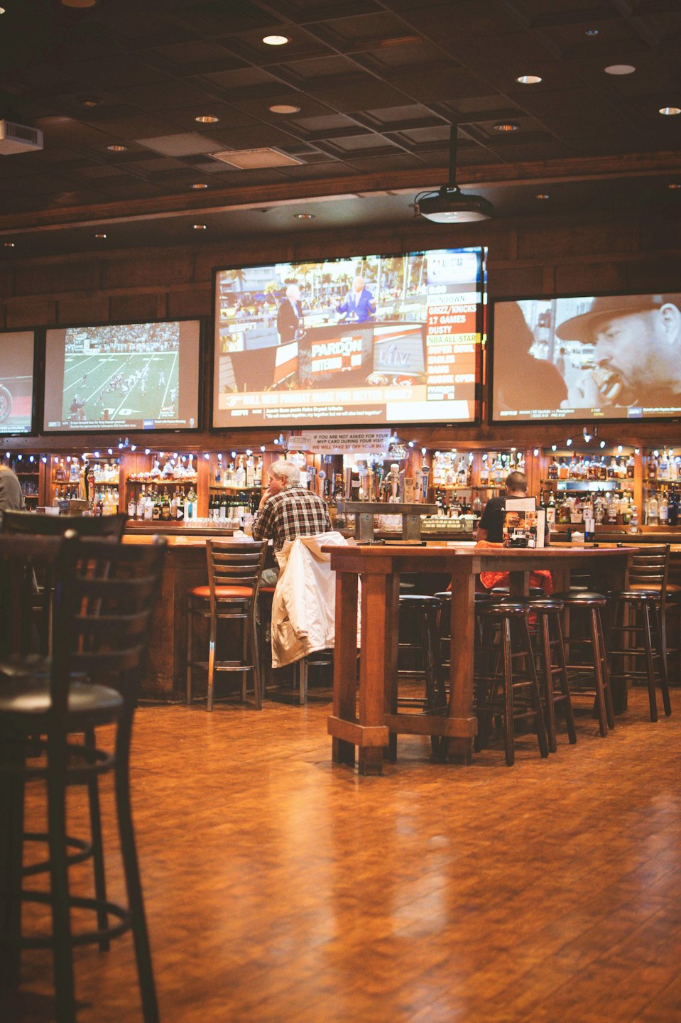 Patrons watch sports on television screens at a wooden bar.