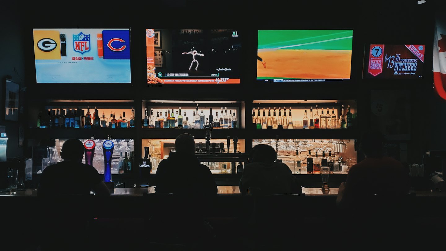 Patrons sitting at a bar counter beneath several television screens.